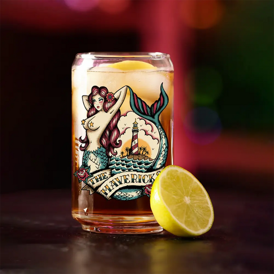 Glass containing a beverage with a mermaid and lighthouse illustration on it, accompanied by a lemon slice.